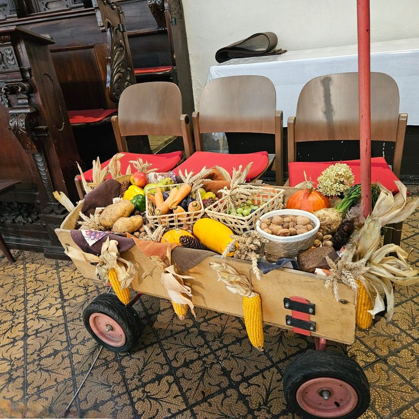 Ein hölzerner Wagen, gefüllt mit verschiedenen Gemüsesorten, Früchten und Nüssen, ist drinnen ausgestellt. Im Hintergrund befinden sich Stühle und ein Tisch.