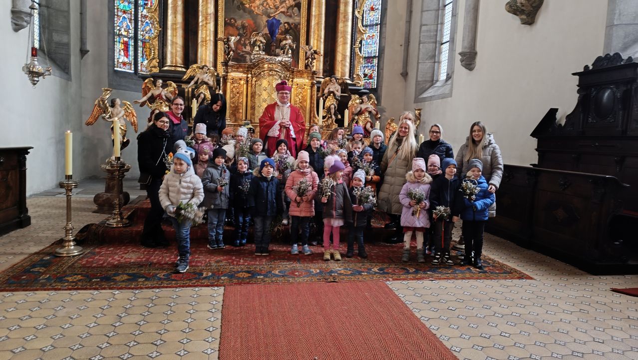 Eine Gruppe von Kindern und Erwachsenen, warm gekleidet, versammelt sich in einer Kirche und hält Blumen. Ein Priester steht hinter ihnen, und Statuen sind im Hintergrund.
