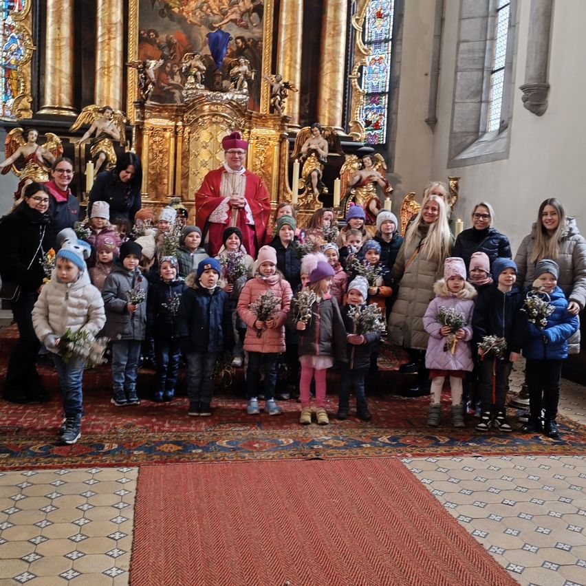 Eine Gruppe von Kindern und Erwachsenen, warm gekleidet, versammelt sich in einer Kirche und hält Blumen. Ein Priester steht hinter ihnen, und Statuen sind im Hintergrund.
