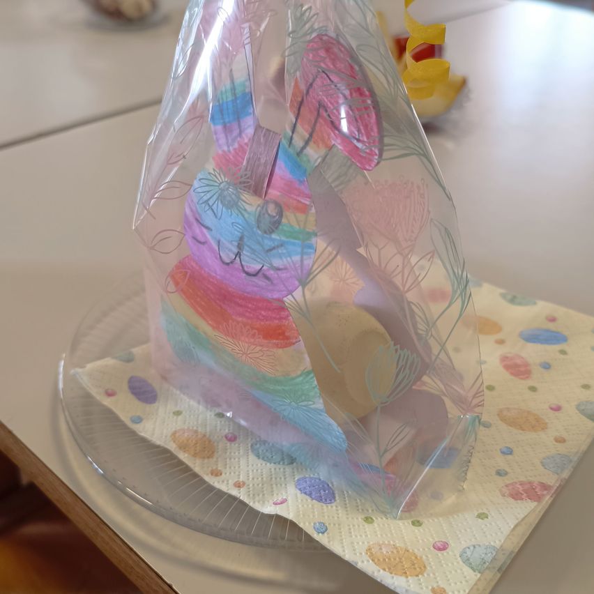 Eine Plastiktüte mit einer bunten Zeichnung eines Hasen liegt auf einer bunten Serviette auf einem klaren Teller. Der Hase ist in Plastik eingewickelt und hat eine farbenfrohe Zeichnung darauf.
