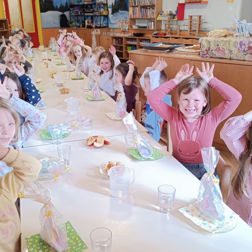 Kinder sitzen an einem langen Tisch mit Essen und Getränken, einige mit erhobenen Händen, in einem Raum mit Bücherregalen und einer Wandmalerei.
