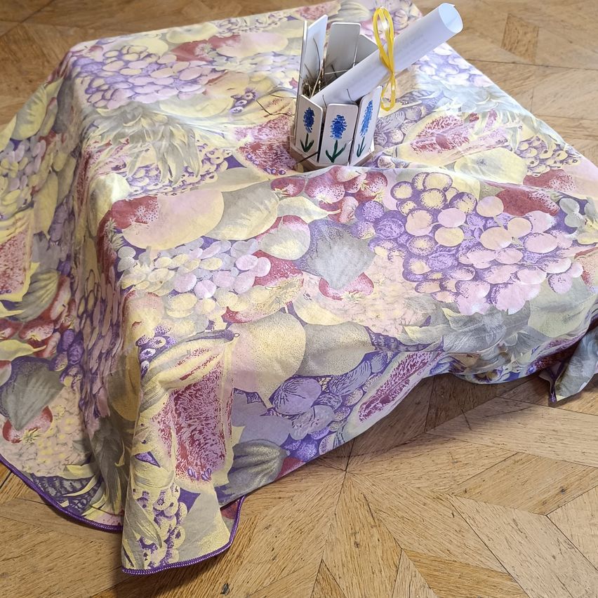 Ein Tischtuch mit Blumen und Früchten, das einen Tisch bedeckt, mit einer kleinen Schachtel, die ein aufgerolltes Papier und ein gelbes Band oben enthält.