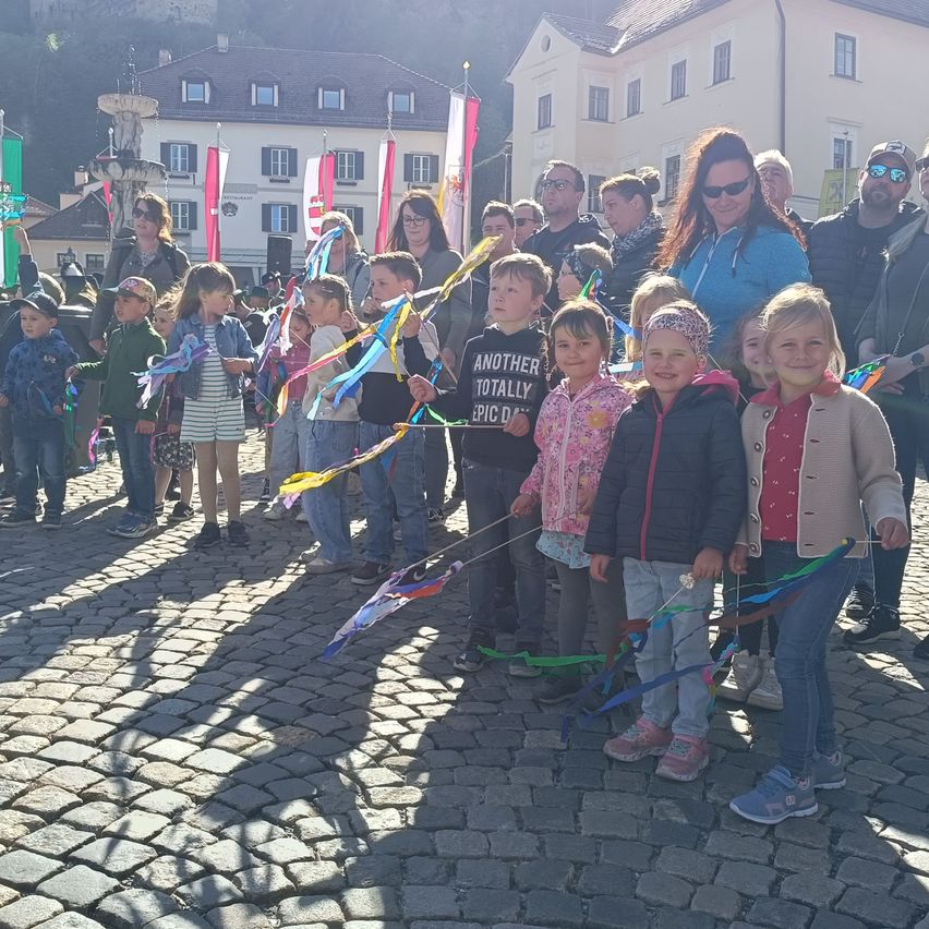 Eine Gruppe von Kindern und Erwachsenen steht in einer Reihe auf einer Kopfsteinpflasterstraße und hält bunte Bänder in der Hand. Sie scheinen an einer Veranstaltung teilzunehmen. Hinter ihnen stehen Gebäude und ein Springbrunnen.