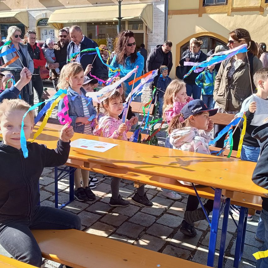 Kinder halten bunte Bänder und sitzen um einen Tisch draußen. Einige Erwachsene stehen hinter ihnen, andere beobachten. Die Kinder schauen auf die Bänder, und einige halten sie hoch.