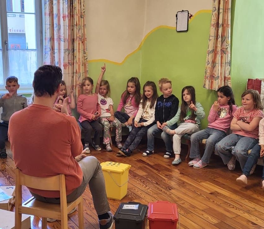 Ein Mann sitzt auf einem Stuhl und leitet eine Gruppe von Kindern, die in einer Reihe sitzen. Bunte Behälter stehen auf dem Boden.