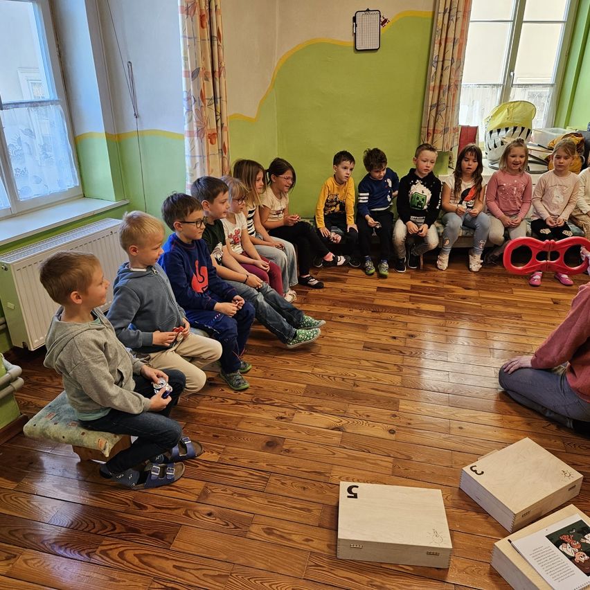 Eine Gruppe von Kindern sitzt in einem Raum und schaut interessiert auf etwas. Einige tragen Brillen und Turnschuhe. Eine Person sitzt auf dem Boden rechts. Holzkisten liegen auf dem Boden.