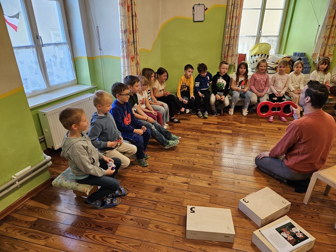 Eine Gruppe von Kindern sitzt in einem Raum und schaut interessiert auf etwas. Einige tragen Brillen und Turnschuhe. Eine Person sitzt auf dem Boden rechts. Holzkisten liegen auf dem Boden.