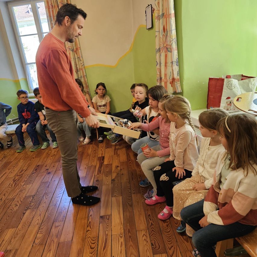 Ein Mann verteilt Spielzeug an Kinder, die in einem Kreis sitzen. Die Kinder schauen erwartungsvoll zu und warten auf ihren Zug.