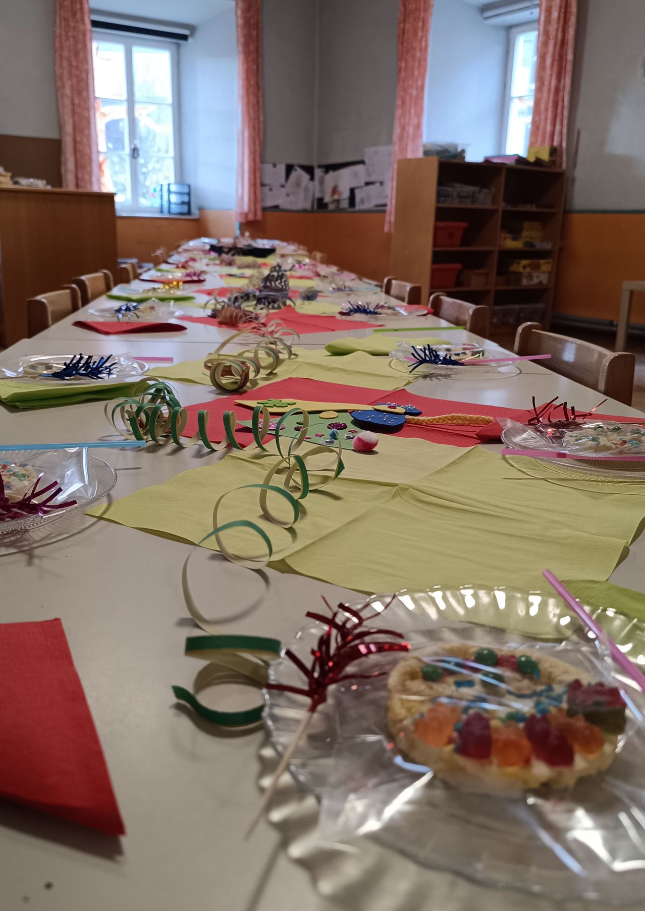 Ein langer Tisch ist mit bunten Dekorationen, Tellern und Servietten gedeckt, einschließlich eines Desserts auf einem Teller mit Gummibonbons. Hinter dem Tisch steht ein Holzregal mit verschiedenen Gegenständen.