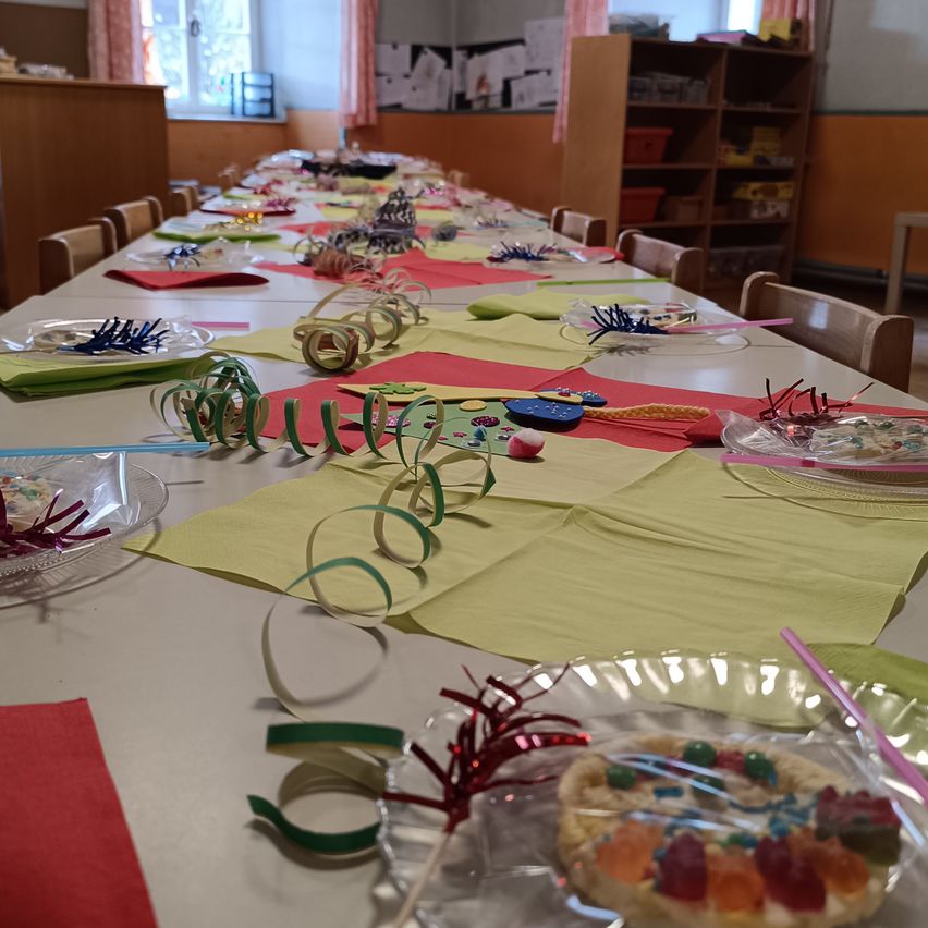 Ein langer Tisch ist mit bunten Dekorationen, Tellern und Servietten gedeckt, einschließlich eines Desserts auf einem Teller mit Gummibonbons. Hinter dem Tisch steht ein Holzregal mit verschiedenen Gegenständen.