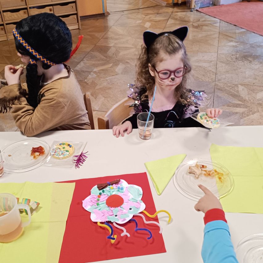 Zwei Kinder sitzen am Tisch und essen. Das Kind auf der linken Seite trägt eine Kopfbedeckung und ein braunes Kostüm. Das Kind auf der rechten Seite trägt Katzenohren und eine Brille. Auf dem Tisch stehen Teller mit Essen, ein Getränk in einem Glas und Servietten.