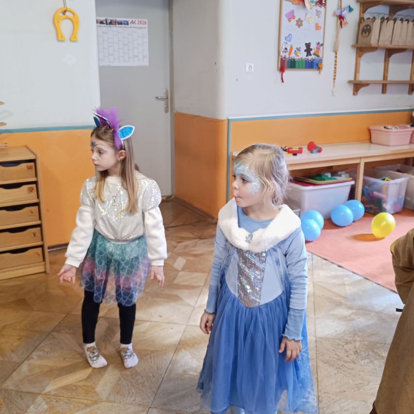 Zwei kleine Mädchen stehen in einem Raum. Ein Mädchen ist als Einhorn und das andere als Fee verkleidet. Sie stehen vor einer Tür mit Luftballons.