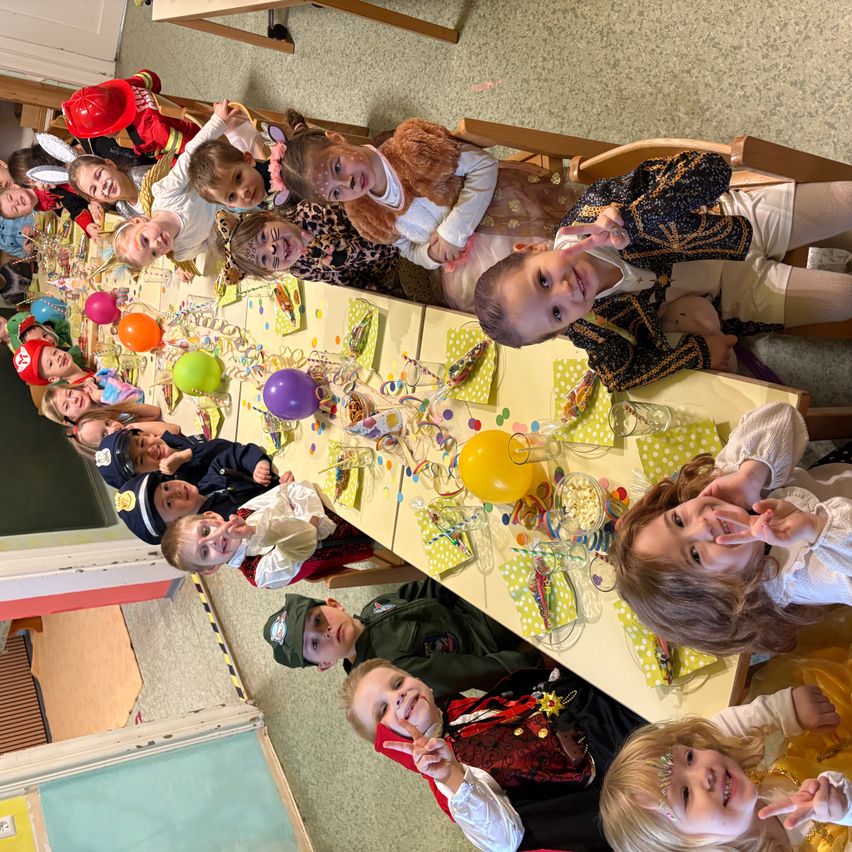 Eine Gruppe von Kindern in Kostümen sitzt an einem langen Tisch, geschmückt mit Luftballons und Dekorationen. Sie scheinen sich auf einem festlichen Zusammentreffen zu befinden.