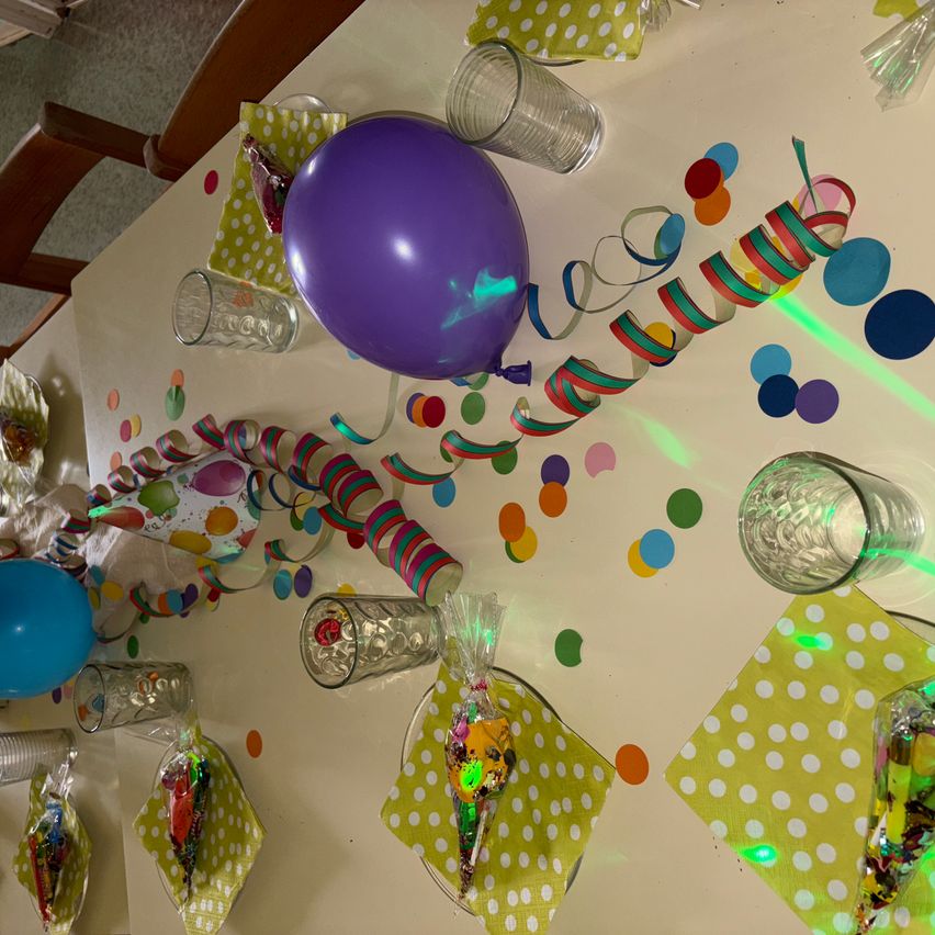 Eine festliche Tischdekoration mit bunten Luftballons, Konfetti und Partydekorationen. Klare Gläser und Süßigkeiten sind auf grünen Punktenservietten angeordnet. Ein lila Luftballon sticht besonders hervor.
