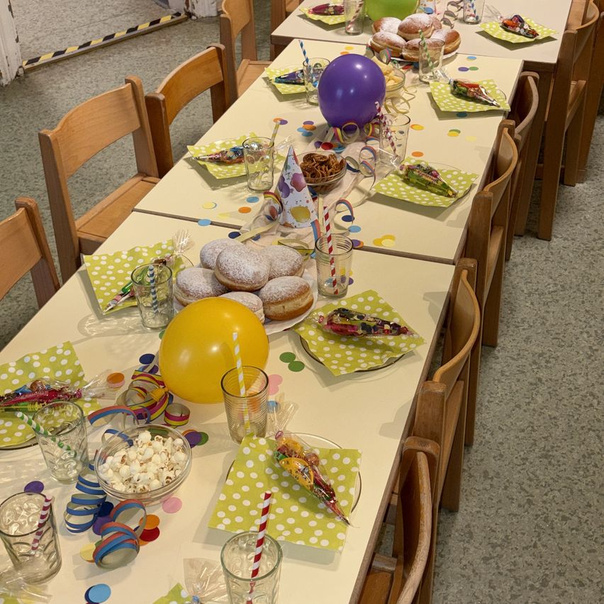 Ein langer Tisch ist mit Luftballons, Konfetti und Snacks dekoriert. Teller mit Donuts und Popcorn sind arrangiert, und farbenfrohe Papierservietten liegen auf jedem Stuhl.