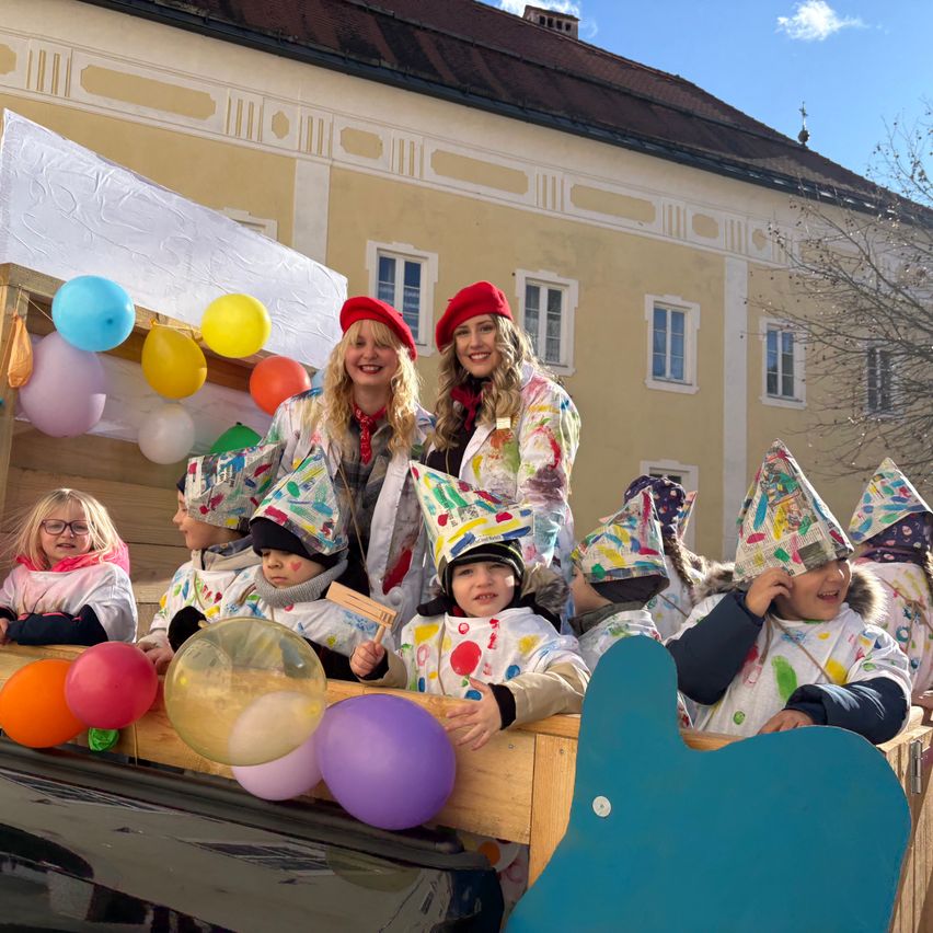 Zwei Frauen und mehrere Kinder, gekleidet in verspielte Kostüme und mit bunten Partyhüten, sitzen auf einem mit Luftballons geschmückten Wagen bei einer Parade.