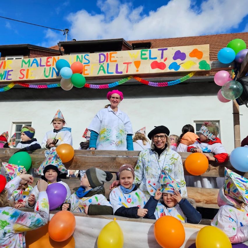 Eine Gruppe von Kindern in bunt bemalten Klamotten und Mützen sitzt auf einer Holzbank und hält Luftballons. Eine Frau steht hinter ihnen, ebenfalls in einer bemalten Kleidung und lächelt. Ein Banner mit einer Regenbogenfarbenpalette und einer Botschaft hängt über ihnen.