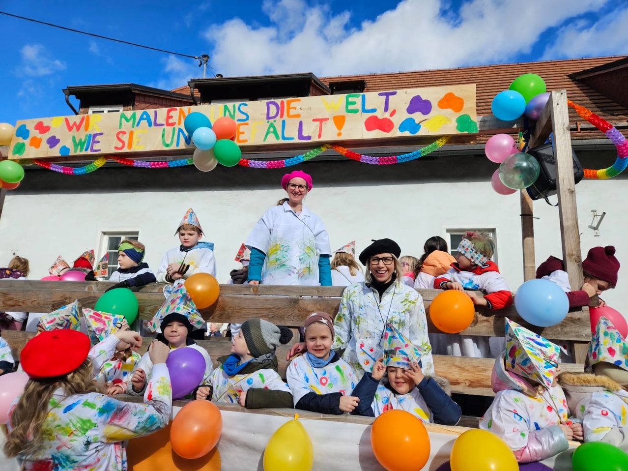 Eine Gruppe von Kindern in bunt bemalten Klamotten und Mützen sitzt auf einer Holzbank und hält Luftballons. Eine Frau steht hinter ihnen, ebenfalls in einer bemalten Kleidung und lächelt. Ein Banner mit einer Regenbogenfarbenpalette und einer Botschaft hängt über ihnen.