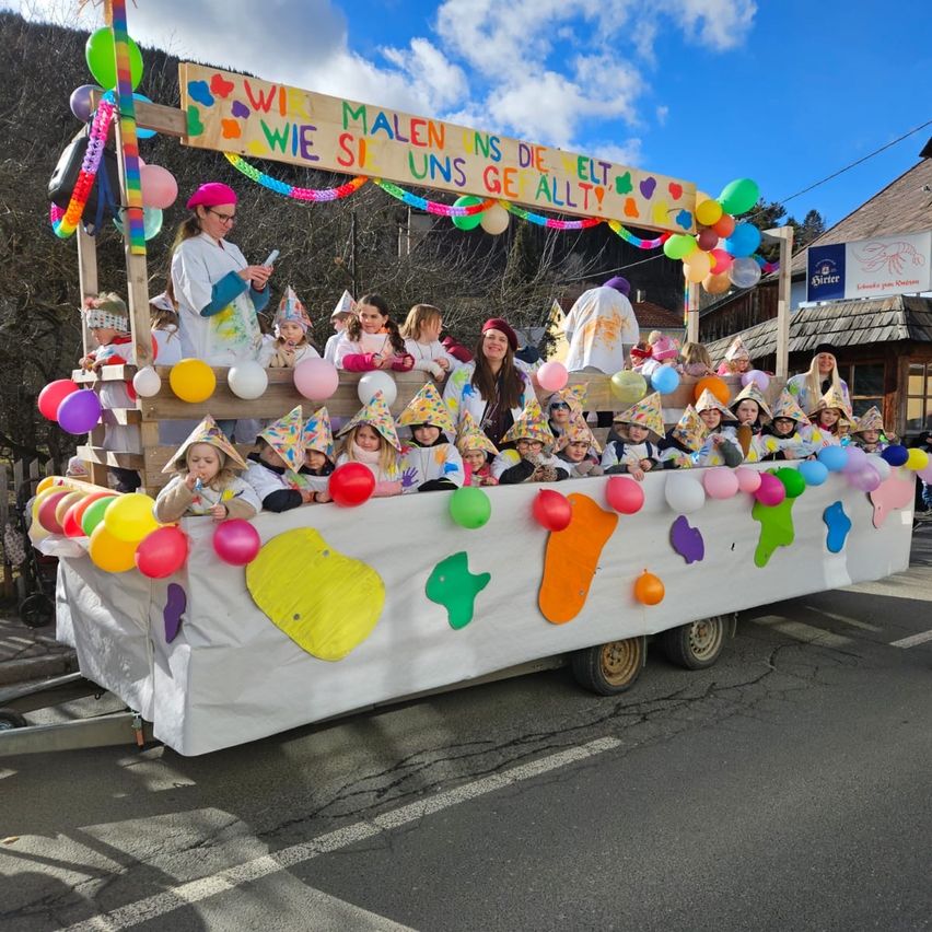 Ein Umzugswagen mit einem Schild, das 'Wir sind alle wie Kinder' besagt, Kinder in Kostümen, bunten Luftballons und Dekorationen an einem sonnigen Tag.