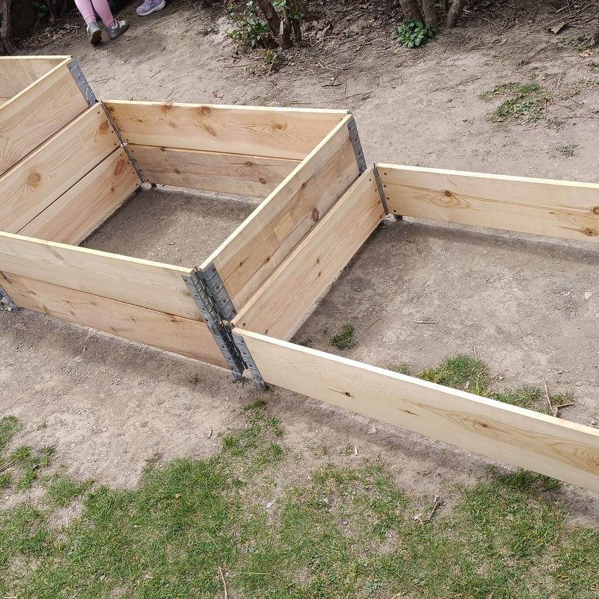 Ein hölzernes Hochbeet mit mehreren Abschnitten, auf einer sandigen Oberfläche mit etwas Gras.