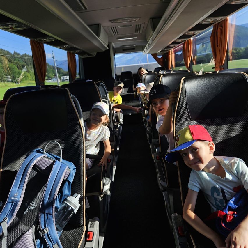 Im Inneren eines Busses sitzen Kinder in schwarzen Sitzen mit Rucksäcken. Ein Junge trägt eine bunte Kappe. Grüne Landschaft ist durch die Fenster sichtbar.