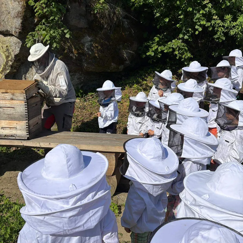 Ein Imker in Schutzkleidung steht vor einem Bienenstock, während mehrere Personen, möglicherweise Kinder, ebenfalls Schutzanzüge und -hüte tragen.