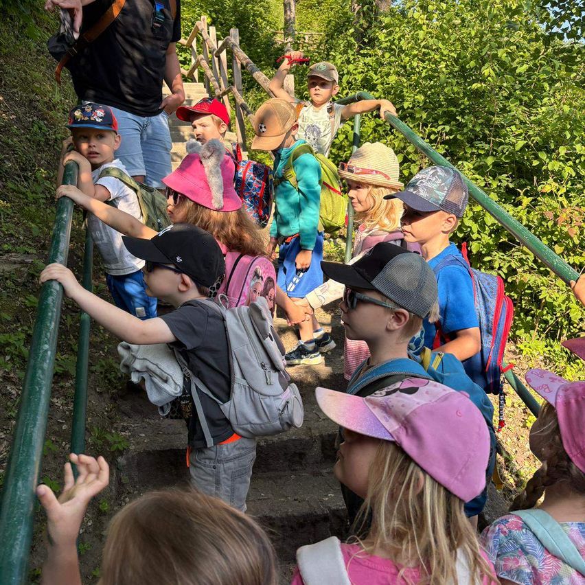 Eine Gruppe von Kindern, einige mit Hüten und Brillen, gehen eine grüne Treppe hinauf und halten sich am Handlauf fest. Sie haben Rucksäcke auf dem Rücken, und an der Seite befindet sich ein grünes Geländer.