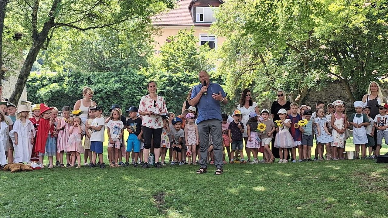 Ein Mann mit Mikrofon steht vor einer Gruppe von Kindern mit Blumen in einem Park.