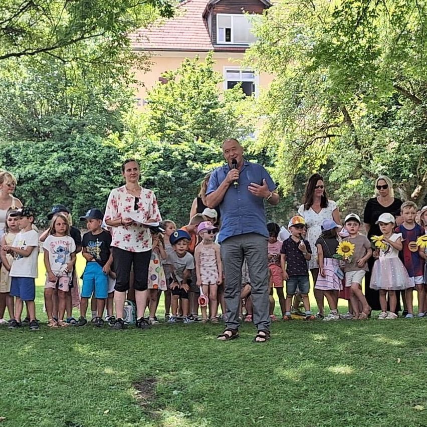 Ein Mann mit Mikrofon steht vor einer Gruppe von Kindern mit Blumen in einem Park.