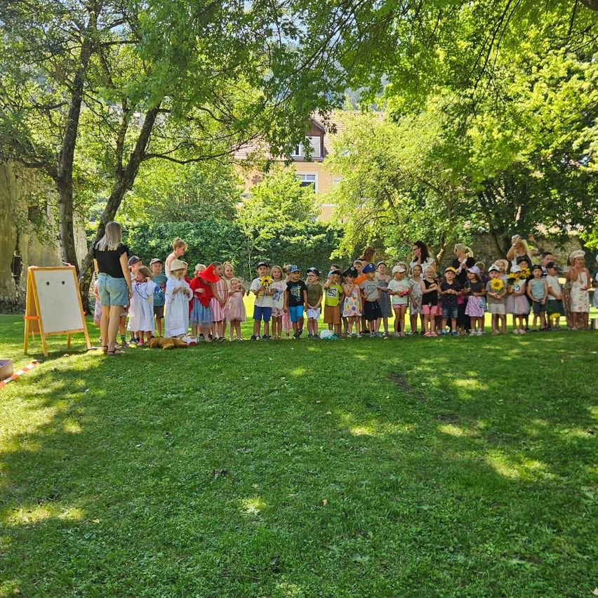 Eine Gruppe von Kindern und Erwachsenen steht an einem sonnigen Tag in einem Park, einige halten Sonnenblumen. Bäume und ein Gebäude sind im Hintergrund.