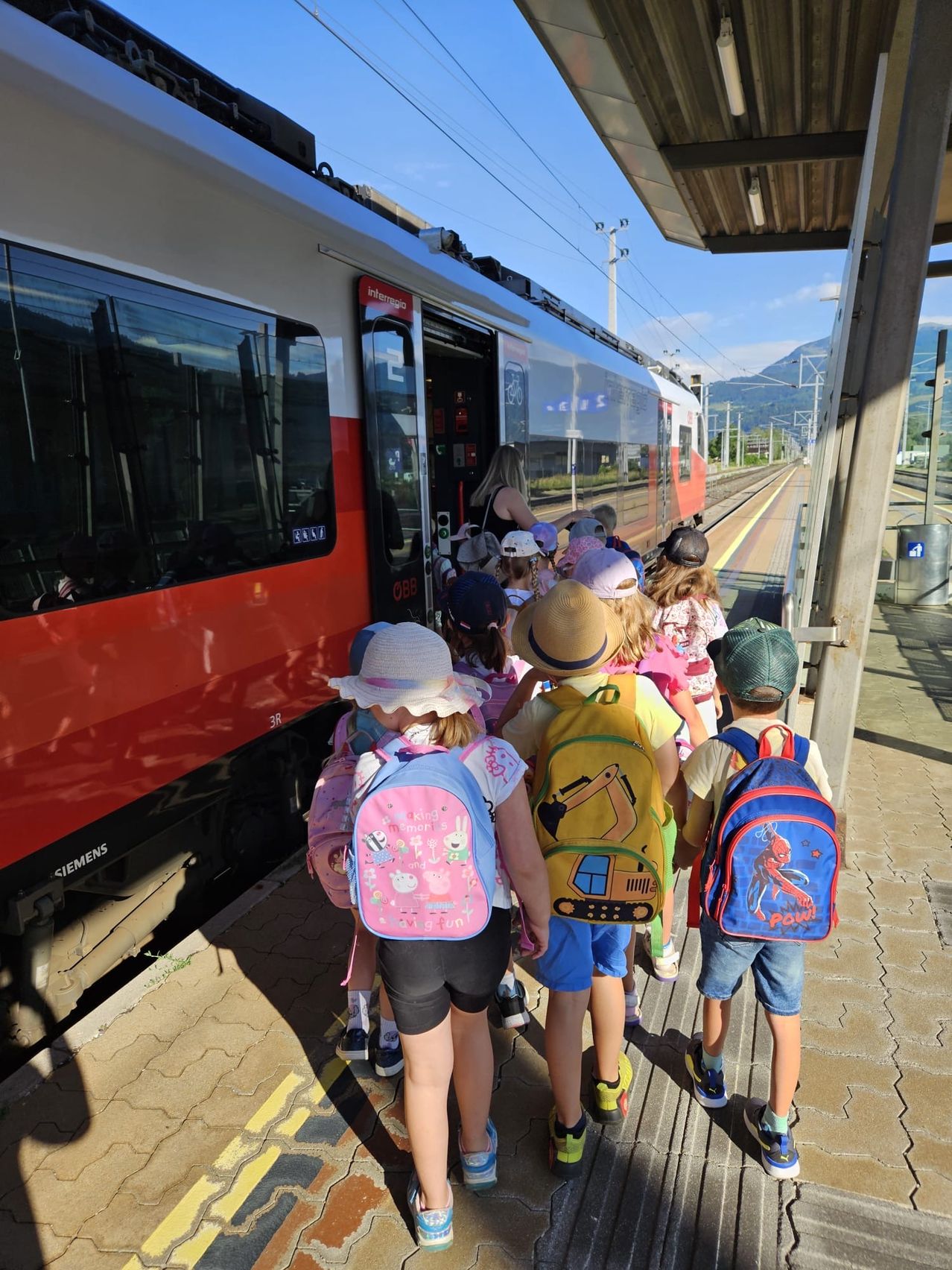 Eine Gruppe von Kindern steigt an einem sonnigen Tag in einen Zug. Sie tragen Hüte und Rucksäcke und werden von einer Frau begleitet. Der Zug ist rot und weiß.