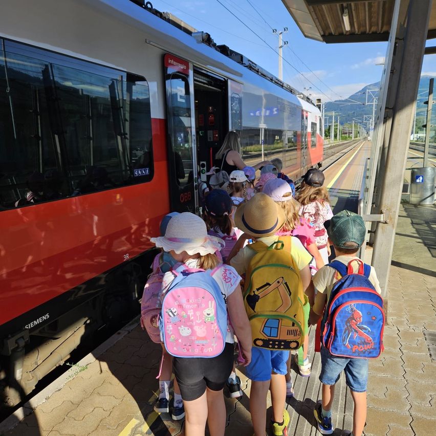 Eine Gruppe von Kindern steigt an einem sonnigen Tag in einen Zug. Sie tragen Hüte und Rucksäcke und werden von einer Frau begleitet. Der Zug ist rot und weiß.