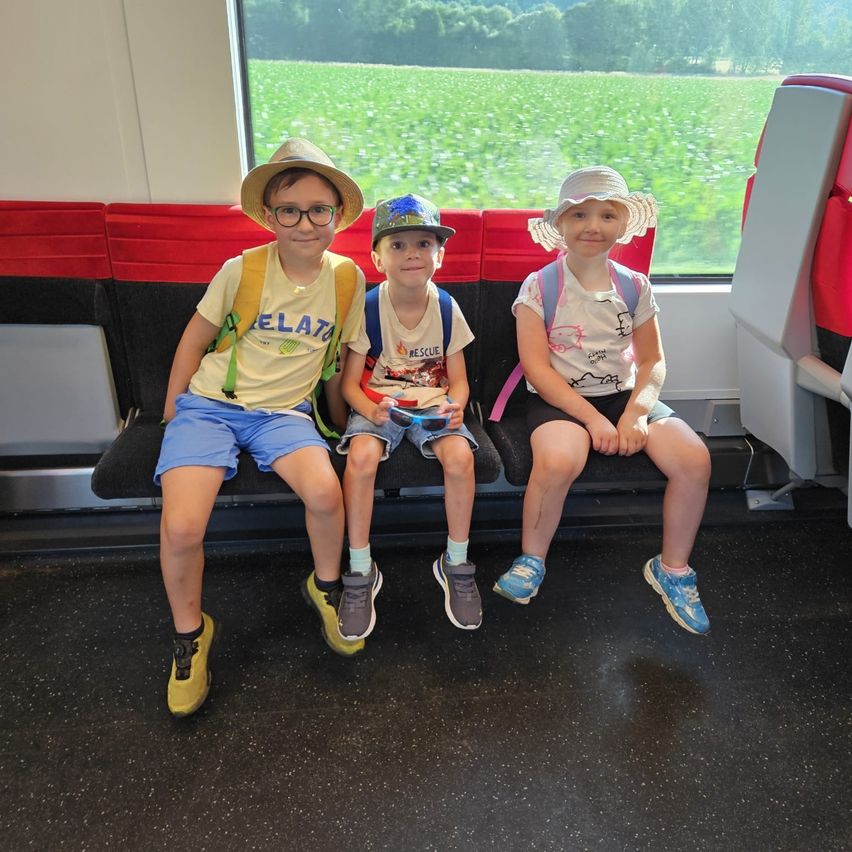 Drei Kinder sitzen auf einem Zug, lächelnd, mit Blick auf ein Feld durch das Fenster. Sie tragen Hüte, Brillen und Rucksäcke.