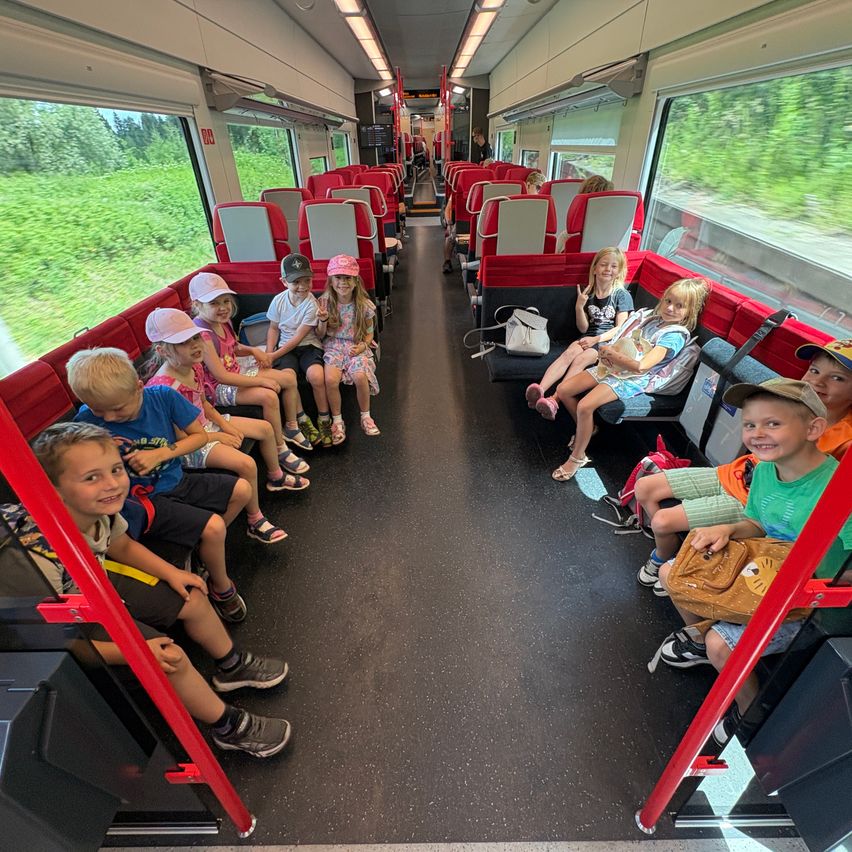 Eine Gruppe von Kindern sitzt mit roten Sitzen auf einem Zug, einige tragen Hüte und schauen aus den Fenstern auf die Grünflächen.