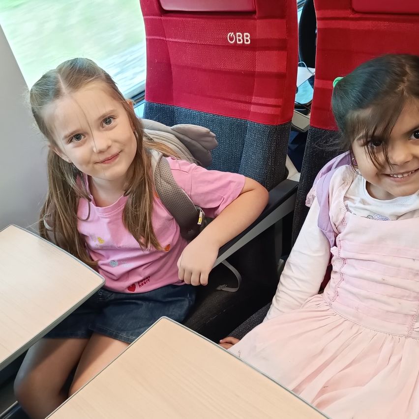 Zwei junge Mädchen sitzen nebeneinander in einem Zug und lächeln. Das Mädchen auf der linken Seite trägt ein rosa Shirt und eine Jeansshorts, während das Mädchen auf der rechten Seite ein rosa Kleid trägt. Die Sitze sind rot und grau, mit einem Tisch vor ihnen. Das Fenster zeigt einen verschwommenen Blick nach draußen.