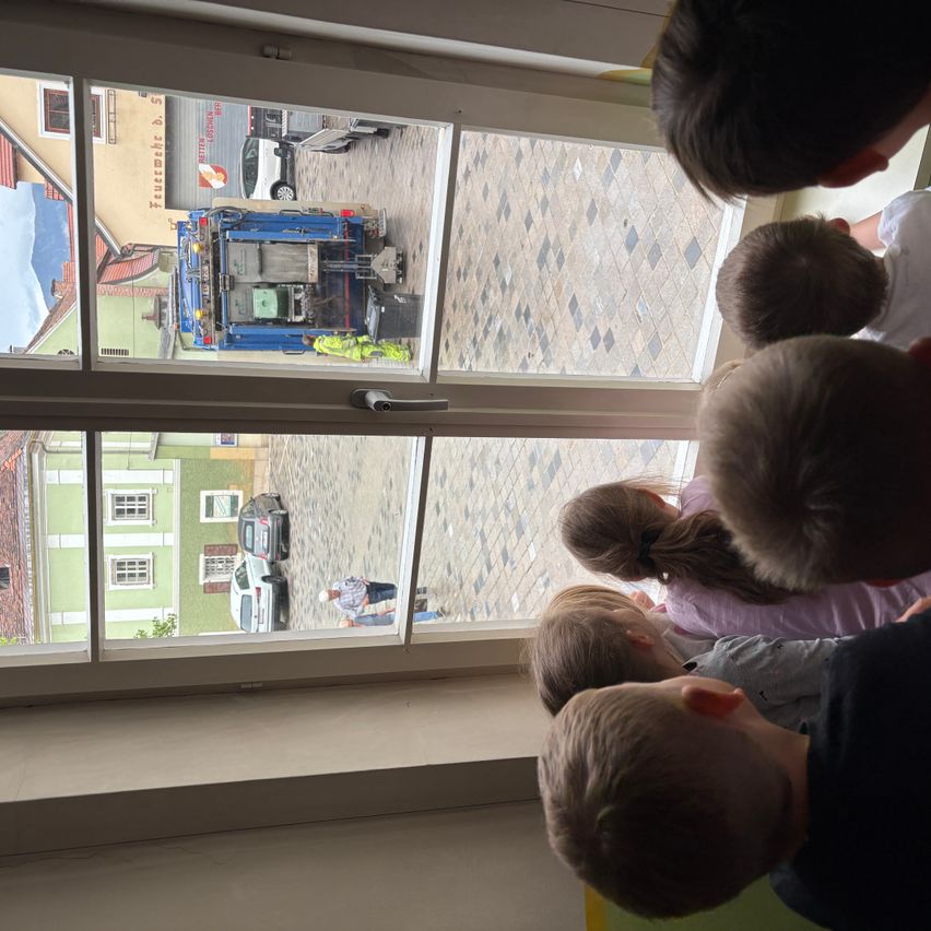 Eine Gruppe von Kindern beobachtet einen Müllwagen durch ein Fenster. Der Wagen steht auf der Straße, und eine Person steht in der Nähe.
