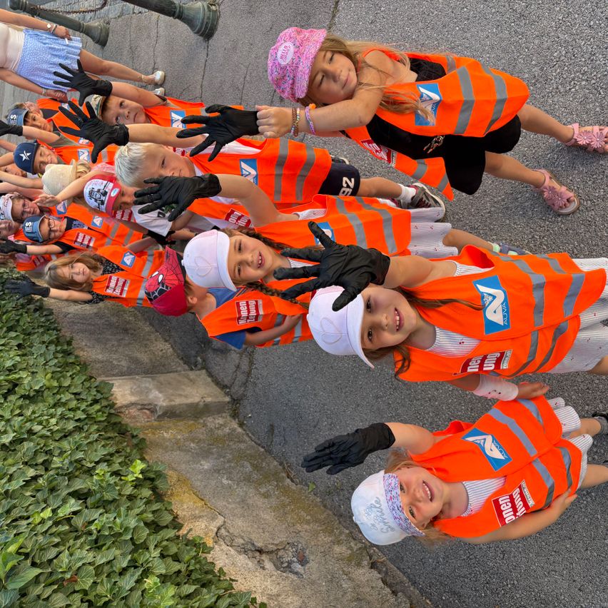 Eine Gruppe von Kindern in orangefarbenen Westen und Handschuhen, die lächelnd für ein Foto auf einer Straße posieren.