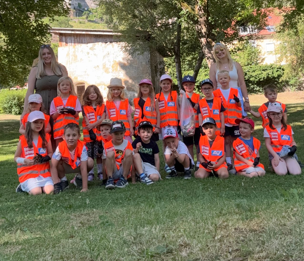 Eine Gruppe von Kindern und zwei Erwachsenen in orangenen Westen posiert für ein Foto im Park. Bäume und ein Gebäude sind im Hintergrund zu sehen.