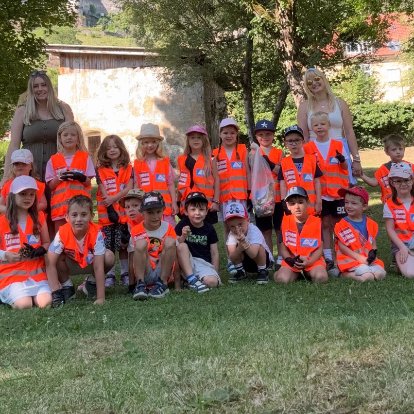 Eine Gruppe von Kindern und zwei Erwachsenen in orangenen Westen posiert für ein Foto im Park. Bäume und ein Gebäude sind im Hintergrund zu sehen.