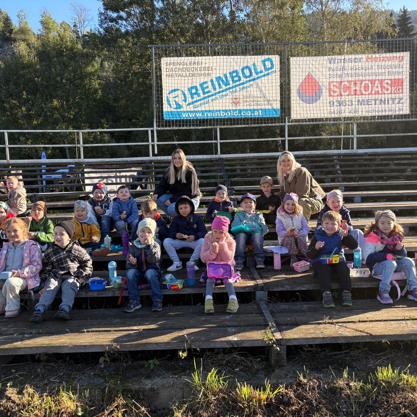 Eine Gruppe von Kindern und zwei Frauen sitzen auf Tribünen, wahrscheinlich bei einem Sportereignis, mit einem Banner dahinter.