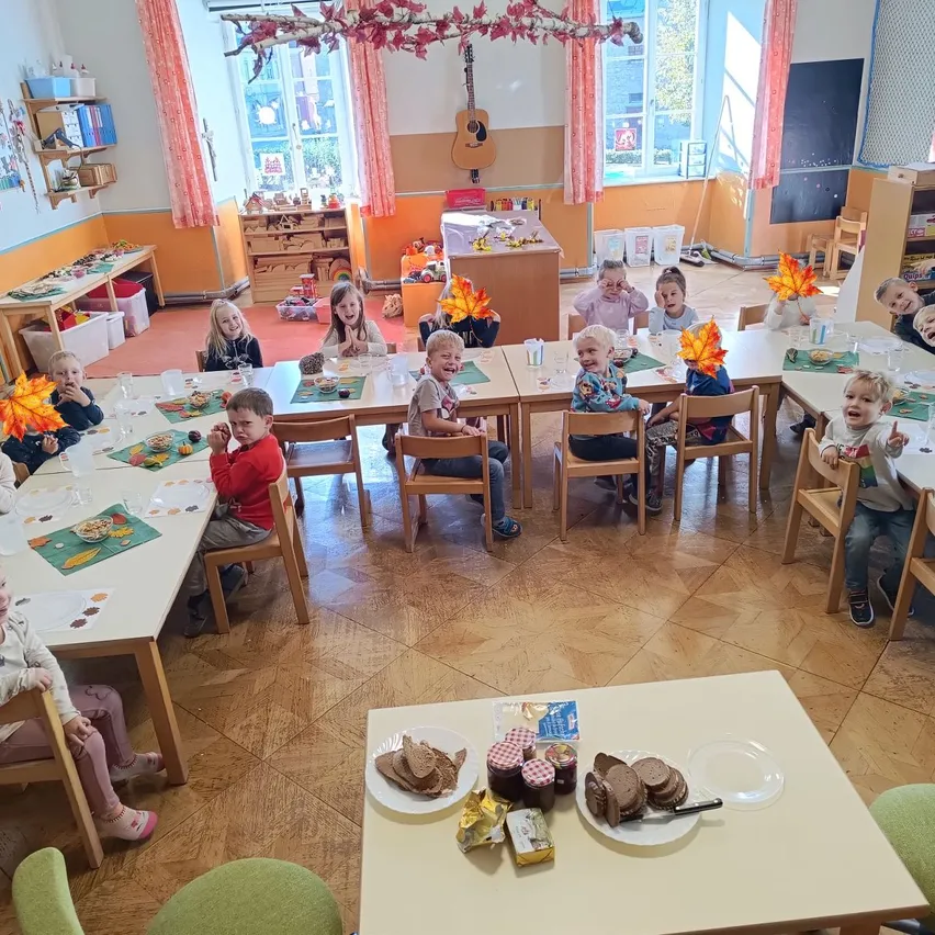 Ein Klassenzimmer mit Kindern, die an Tischen sitzen, essen und lächeln. Der Raum hat Holzböden, weiße Wände und große Fenster.