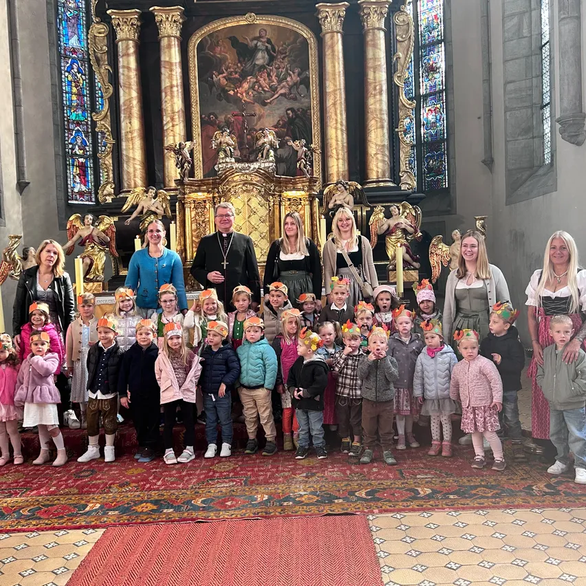 Eine Gruppe von Kindern und Erwachsenen versammelt sich in einer Kirche und trägt festliche Hüte. Die Kirche ist mit einem goldenen Altar und Buntglasfenstern geschmückt.