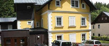 Ein großes gelbes Gebäude mit mehreren Fenstern und einem braunen Mittelteil, vor dem zwei Autos geparkt sind.