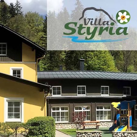 Ein gelbes Gebäude mit Fenstern ist Villa Styria. Es hat ein schwarzes Dach und eine Rutsche. Bäume umgeben es.