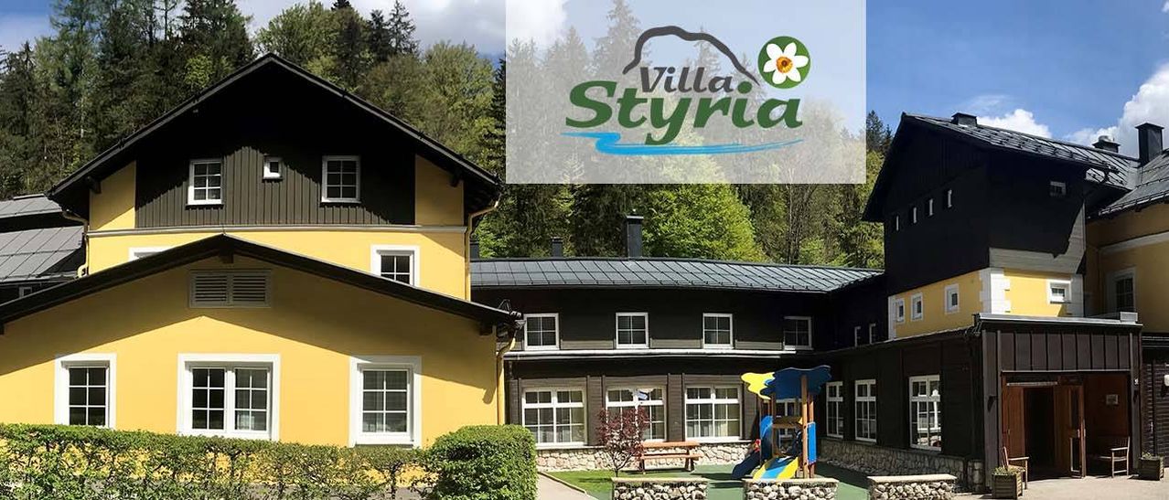 Ein gelbes Gebäude mit Fenstern ist Villa Styria. Es hat ein schwarzes Dach und eine Rutsche. Bäume umgeben es.