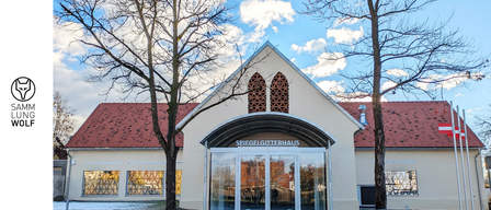 Der Eingang des Spiegelgitterhauses, ein Gebäude mit rotem Dach, verfügt über einen verglasten Eingang mit Überdachung. Über dem Eingang befinden sich zwei Bogenfenster und zwei Bäume flankieren das Gebäude.