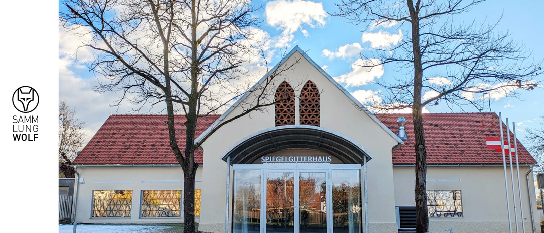 Der Eingang des Spiegelgitterhauses, ein Gebäude mit rotem Dach, verfügt über einen verglasten Eingang mit Überdachung. Über dem Eingang befinden sich zwei Bogenfenster und zwei Bäume flankieren das Gebäude.