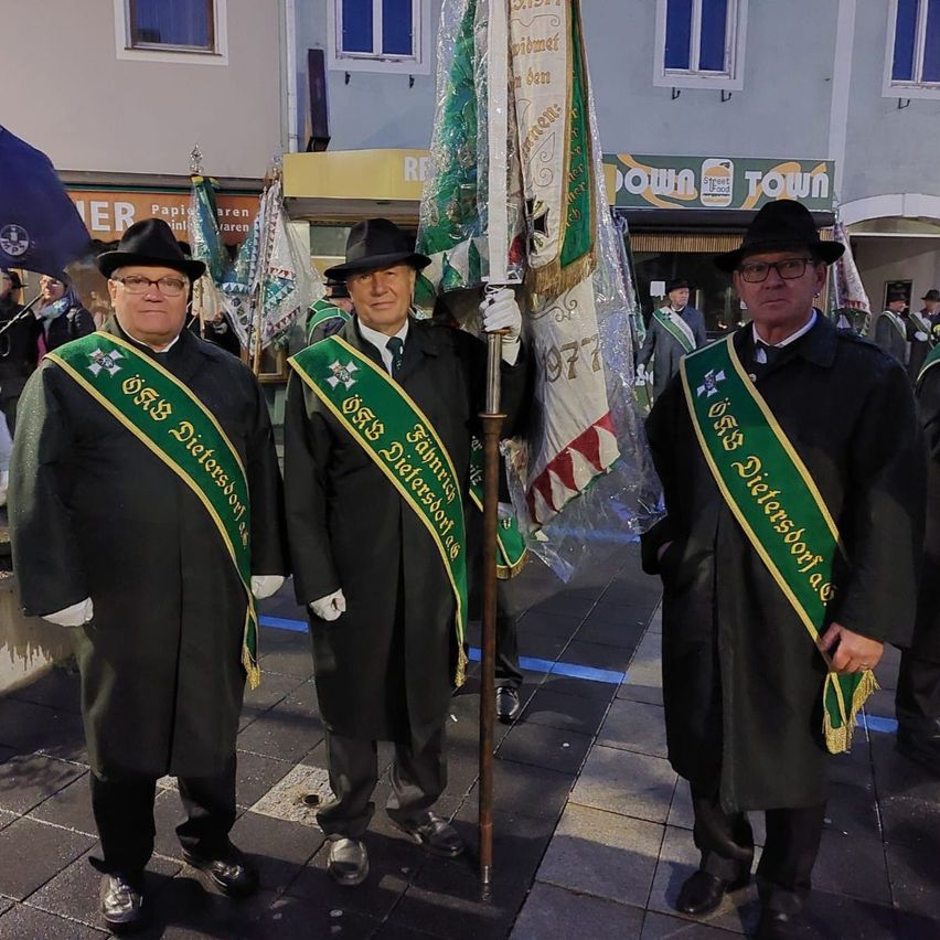Drei Männer in schwarzen Mänteln mit grünen Schärpen stehen zusammen. Sie tragen schwarze Hüte und Brillen. Der Mann in der Mitte hält eine Fahne. Hinter ihnen befinden sich Gebäude mit Schildern und einige Personen im Hintergrund.