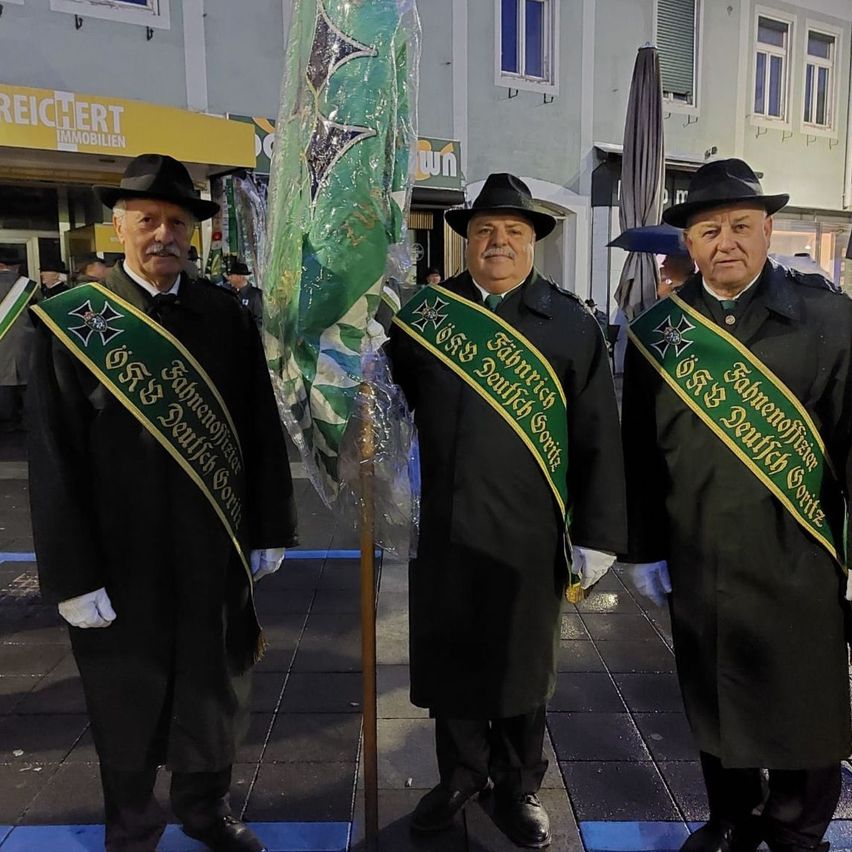 Drei Männer in schwarzen Mänteln mit grünen Schärpen stehen in einer Reihe auf einer Stadtstraße, einer von ihnen hält eine Fahne. Hinter ihnen ist ein Gebäude mit einem Schild, das 'Immobilien' besagt, und ein Regenschirm zu sehen.