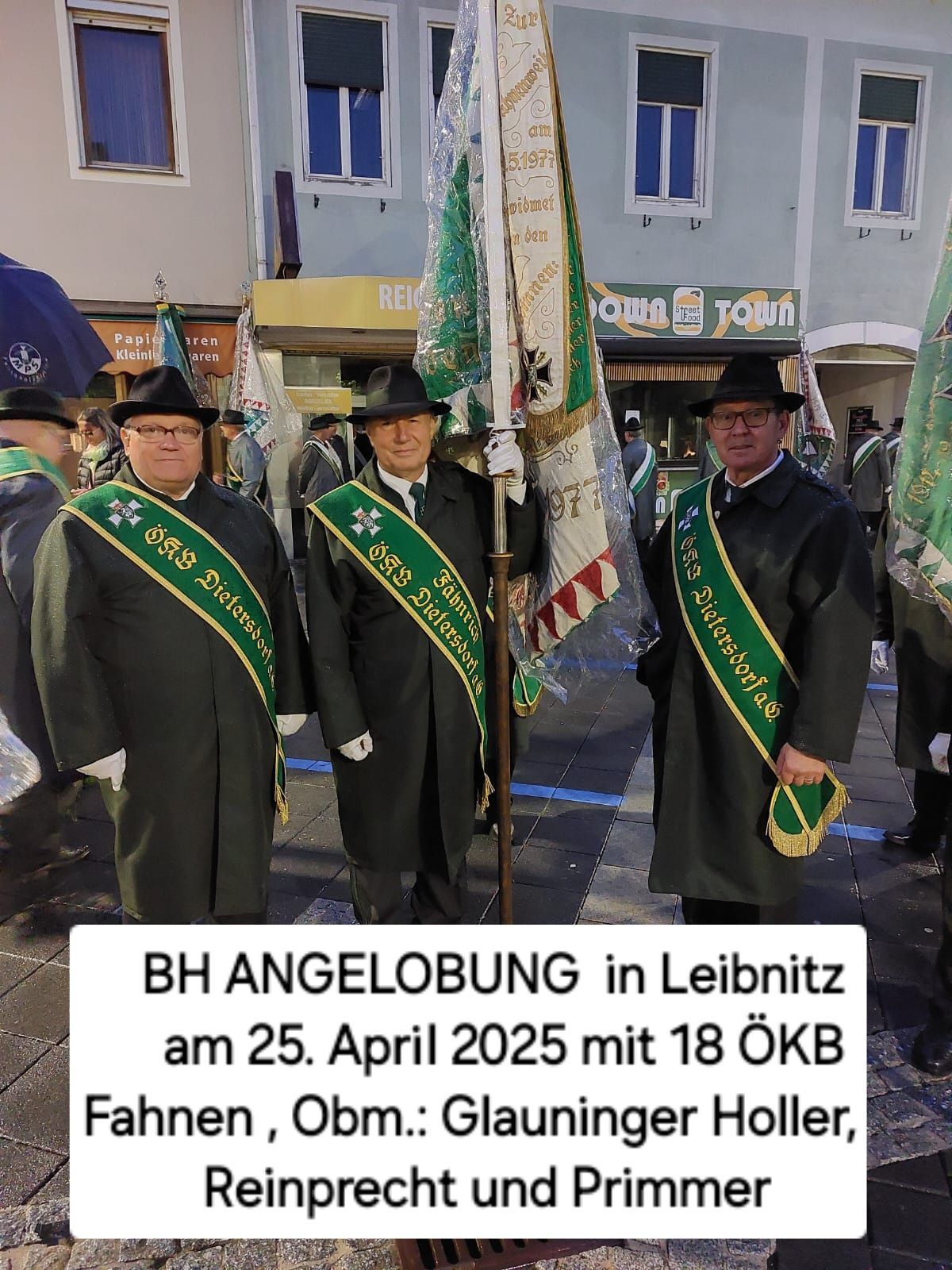 Drei Männer in grünen und goldenen Schärpen, die Hüte tragen und Flaggen halten, stehen vor einem Gebäude mit einer Markise. Hinter ihnen sind weitere Menschen, Gebäude und ein blauer Regenschirm.